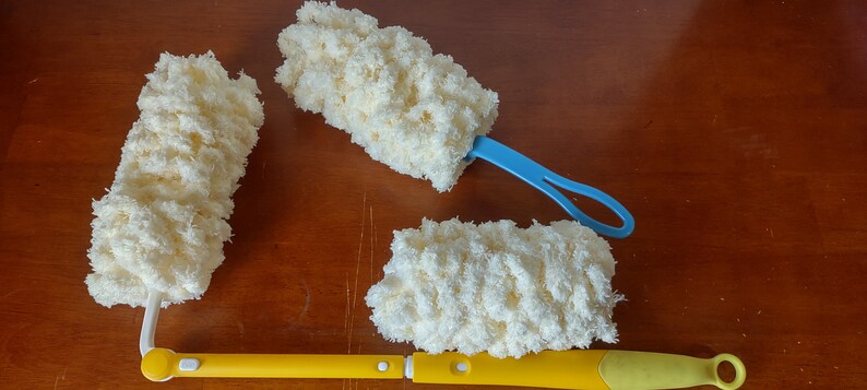 May include: A collection of cleaning dusters with fluffy, cream-colored microfiber heads. One duster has a yellow handle, another has a blue handle, and the third has a white handle. The dusters are arranged on a wooden surface.