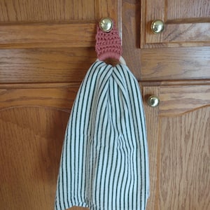 May include: A crocheted towel ring in a light pink colour with a white wooden ring. The ring is hanging on a wooden cabinet door with a white and black striped towel hanging from it.