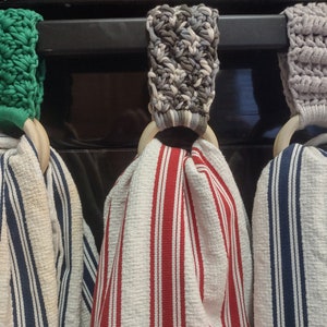 Könnte beinhalten: Drei Küchentuchhalter mit Holzringen. Die Halter sind aus gehäkeltem Stoff in Grün, Grau und Weiß. Die Handtücher sind gestreift in Blau und Weiß, Rot und Weiß und Blau und Weiß.