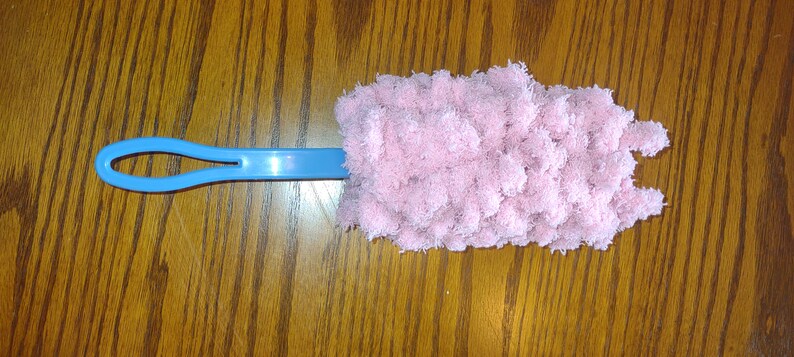 May include: A blue handled duster with a pink fluffy microfiber head. The duster is on a wooden surface.
