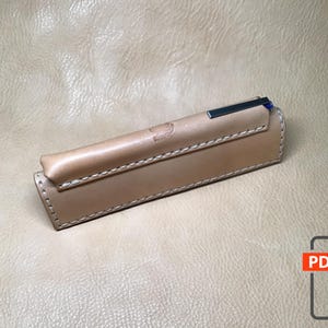 Può includere: Un astuccio per penne in pelle marrone chiaro con un bordo cucito e una penna argentata all'interno. L'astuccio è su una superficie testurizzata marrone chiaro.