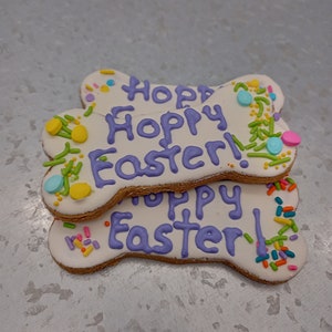 May include: Three bone-shaped dog biscuits decorated with purple icing and colorful sprinkles. The biscuits are stacked on top of each other and say "Happy Easter!" in purple icing.