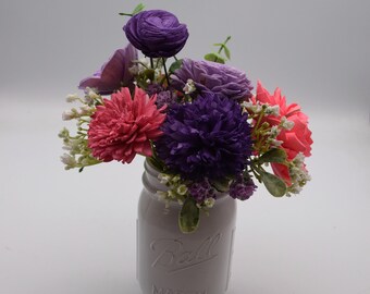 May include: A white Mason jar filled with a colorful floral arrangement. The bouquet features various flowers in shades of purple, pink, and coral, with small white accents. The jar is embossed with the word "Ball" and "MASON".