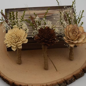 Op de afbeelding: Drie houten bloemenboutonnières met jute stelen. De bloemen zijn lichtbruin, donkerbruin en crèmekleurig. De bloemen zijn gerangschikt op een houten schijf.