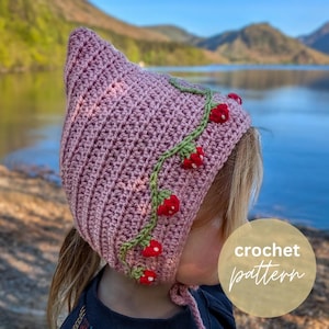 Puede incluir: Un gorro de ganchillo hecho a mano, de color púrpura claro, con una parte superior puntiaguda, adornado con una vid verde y acentos de fresa roja. El gorro tiene una corbata debajo de la barbilla. La imagen también incluye el texto "crochet pattern".
