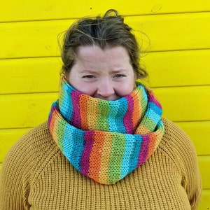 May include: A person wearing a mustard yellow sweater with a colorful striped infinity scarf. The scarf is made of a soft, knitted fabric and features a variety of colors, including blue, green, orange, and pink.