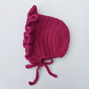 May include: A magenta-colored, crocheted baby bonnet with a ruffled edge and tie closure. The bonnet is handmade with a ribbed texture and a soft, delicate appearance. The ties are long and can be adjusted for a comfortable fit.