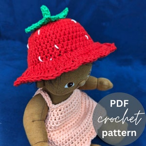 May include: A red crochet strawberry hat with green leaves and white accents. The hat is on a doll with brown skin and a pink crochet dress. The text "PDF crochet pattern" is on a white circle.