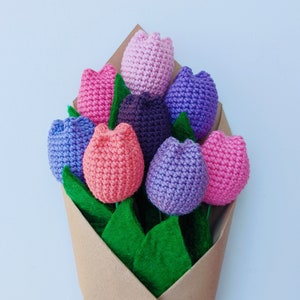 May include: A bouquet of crocheted tulips in shades of pink, purple, and lavender, arranged in a brown paper wrapper with green felt leaves.