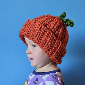 May include: A child wearing a crocheted orange pumpkin hat with a green stem. The hat has a textured, ribbed design.