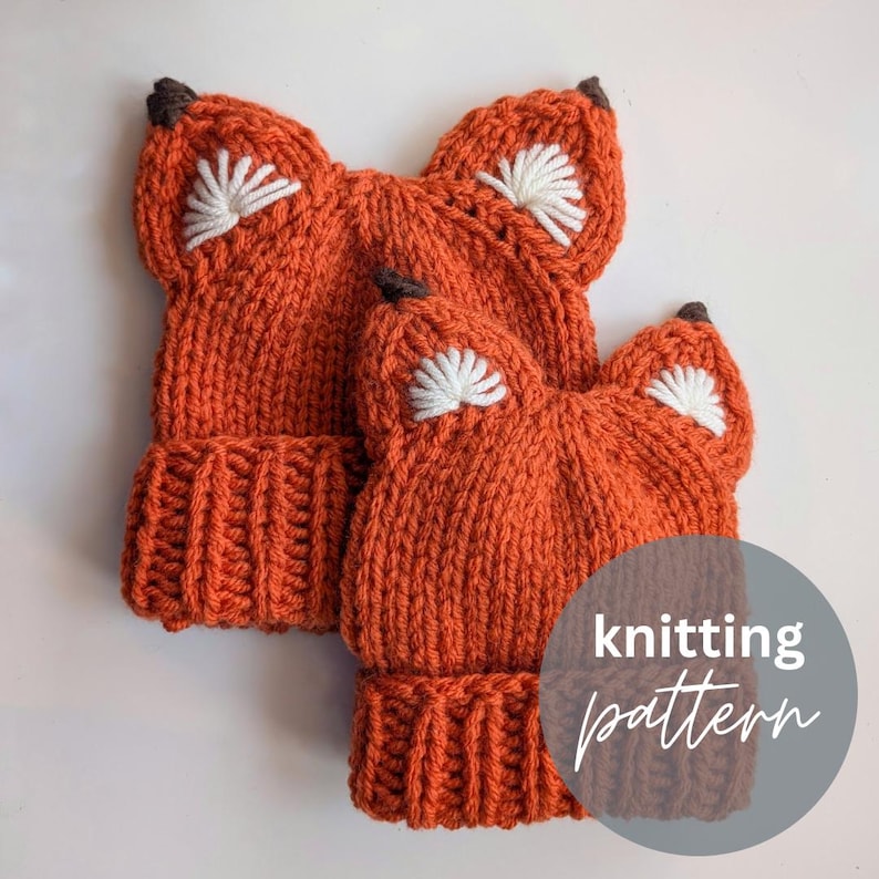 May include: Two orange knitted fox hats with white felt ears and brown felt noses. The hats are on a white surface with the text "knitting pattern" in a gray circle.