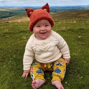 May include: A baby wearing a knitted orange fox hat with white accents, a white knitted sweater, and yellow pants with a colorful animal print. The baby is sitting on a grassy hill with a view of a valley in the background.