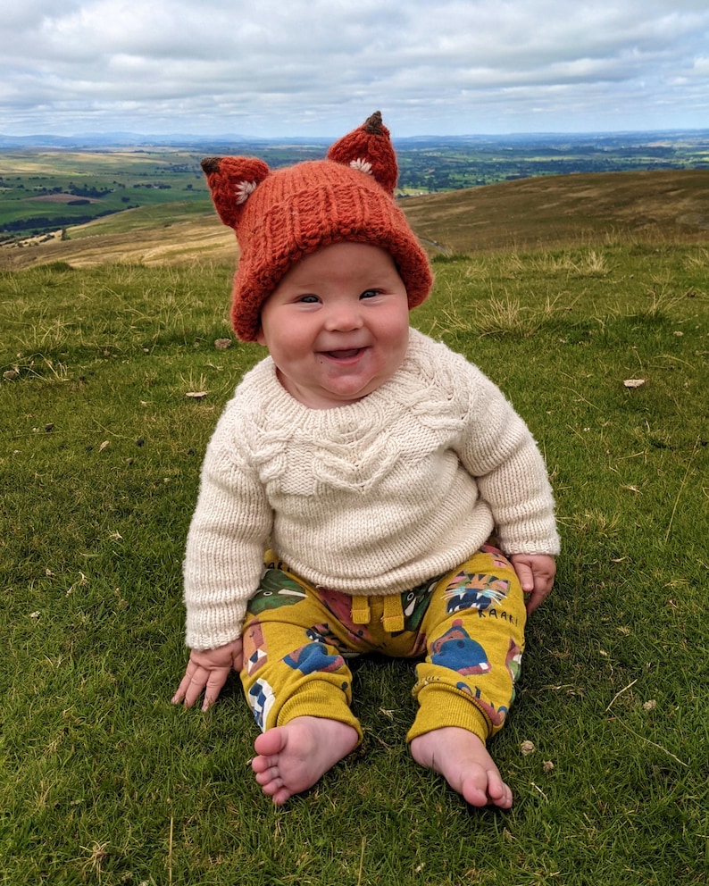 May include: A baby wearing a knitted orange fox hat with white accents, a white knitted sweater, and yellow pants with a colorful animal print. The baby is sitting on a grassy hill with a view of a valley in the background.
