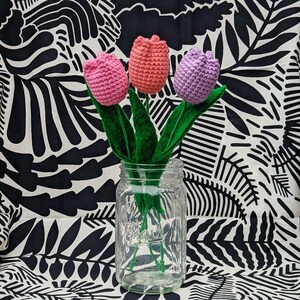 May include: Three crocheted tulips in pink and purple colors in a clear glass jar with a green stem. The jar is sitting on a black and white patterned fabric.