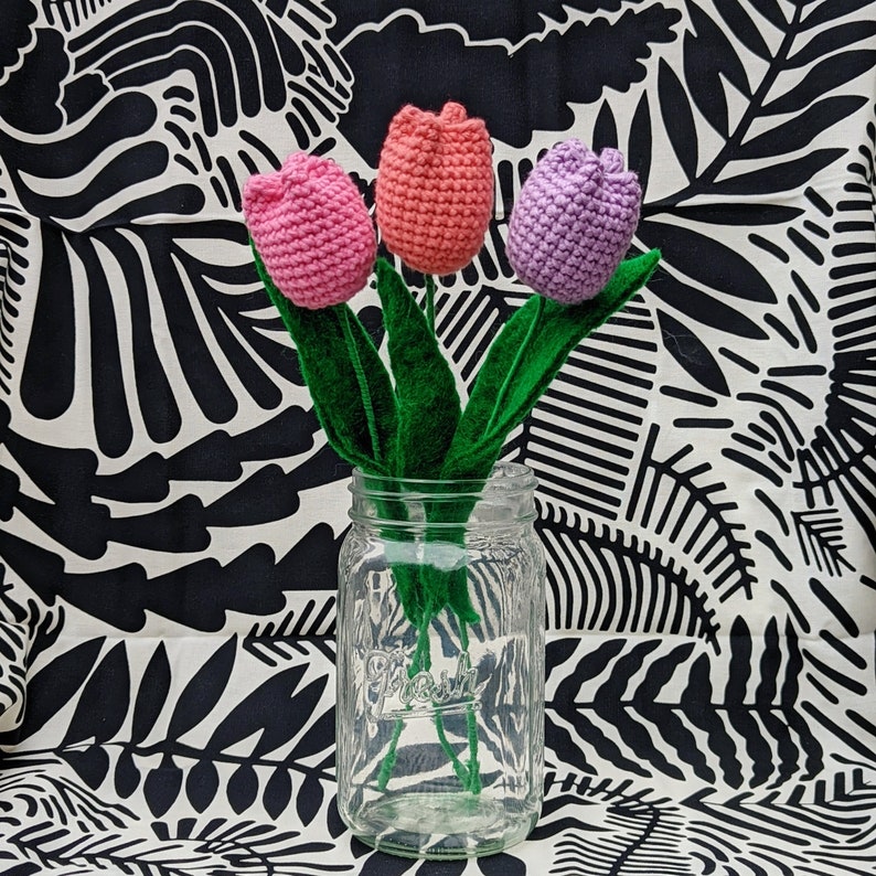 May include: Three crocheted tulips in pink and purple colors in a clear glass jar with a green stem. The jar is sitting on a black and white patterned fabric.