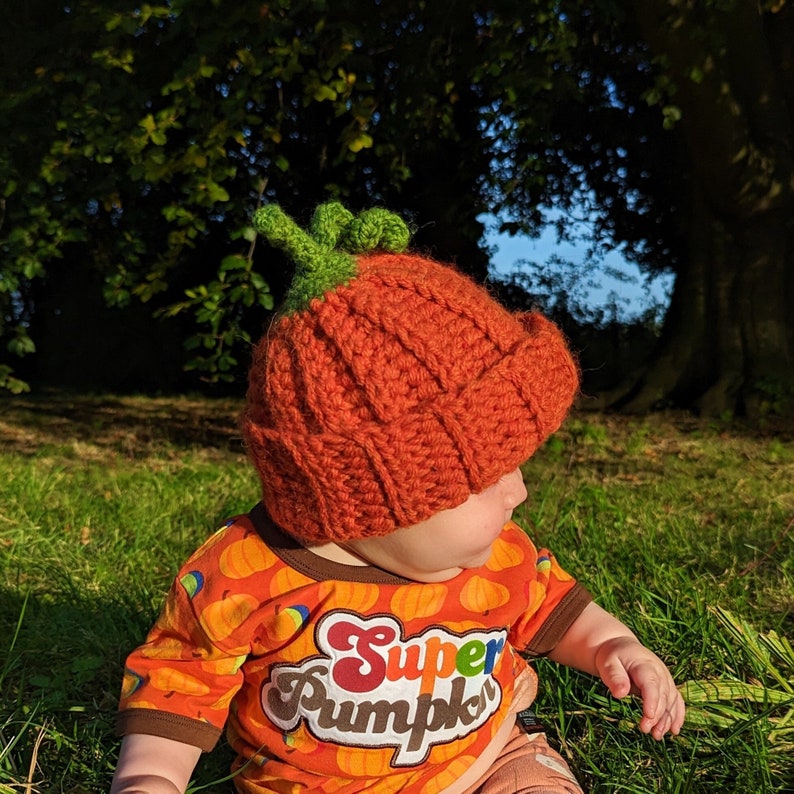 A baby is wearing a crochet orange pumpkin hat with a green stem and a curly leaf.