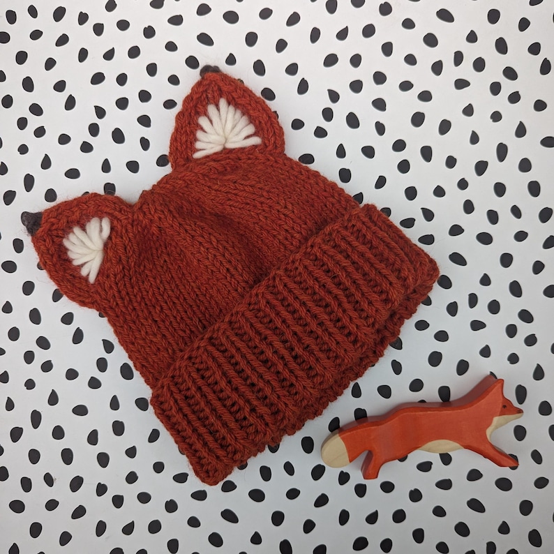 May include: A knitted fox hat in a rust orange color with white felt ears. The hat is on a white background with black polka dots.