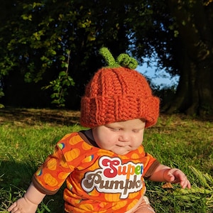 Pode incluir: Uma crian&ccedil;a pequena usando um chap&eacute;u de ab&oacute;bora de croch&ecirc; laranja e marrom com folhas verdes no topo. A crian&ccedil;a est&aacute; usando uma camisa laranja com um gr&aacute;fico branco de "Super Pumpkin". A crian&ccedil;a est&aacute; sentada em uma &aacute;rea gramada.