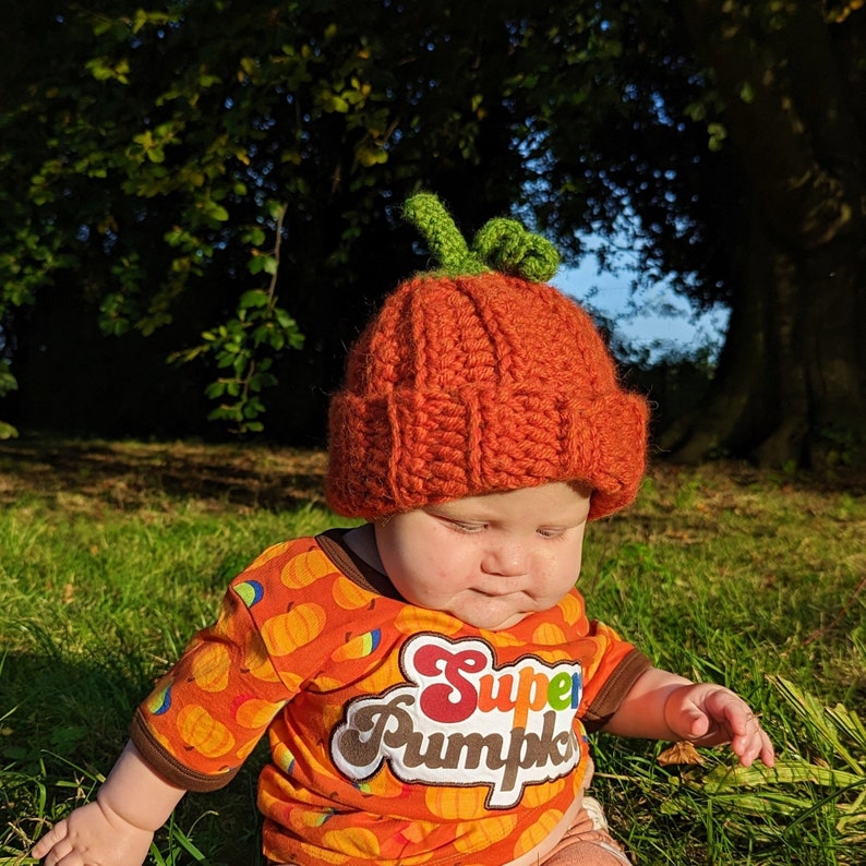 Pode incluir: Uma crian&ccedil;a pequena usando um chap&eacute;u de ab&oacute;bora de croch&ecirc; laranja e marrom com folhas verdes no topo. A crian&ccedil;a est&aacute; usando uma camisa laranja com um gr&aacute;fico branco de "Super Pumpkin". A crian&ccedil;a est&aacute; sentada em uma &aacute;rea gramada.