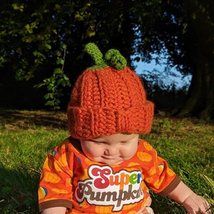 Pode incluir: Uma crian&ccedil;a usando um chap&eacute;u de ab&oacute;bora de croch&ecirc; laranja e uma camisa laranja brilhante com o texto "Super Pumpkin" impresso.