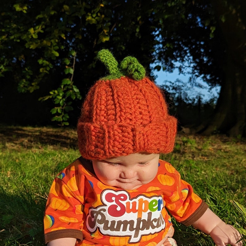 Pode incluir: Uma crian&ccedil;a usando um chap&eacute;u de ab&oacute;bora de croch&ecirc; laranja e uma camisa laranja brilhante com o texto "Super Pumpkin" impresso.