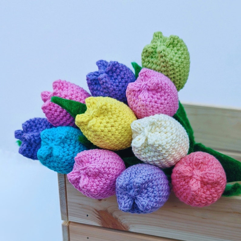 May include: A bouquet of crocheted tulips in a variety of pastel colors, including pink, purple, yellow, blue, and green. The tulips are arranged in a wooden crate.