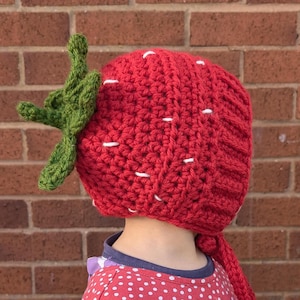 May include: A red crocheted strawberry hat with a green crocheted stem and leaves. The hat has white stitching details and a long tail.