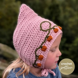 Peut inclure: Un bonnet en crochet rose clair avec un sommet pointu et un motif de vigne décoratif brodé en fil vert foncé, agrémenté de petits accents texturés en forme de citrouille orange sur le côté. Le bonnet possède des liens roses assortis.