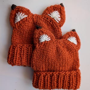May include: Two orange knitted fox hats with white embroidered ears and brown felt noses.