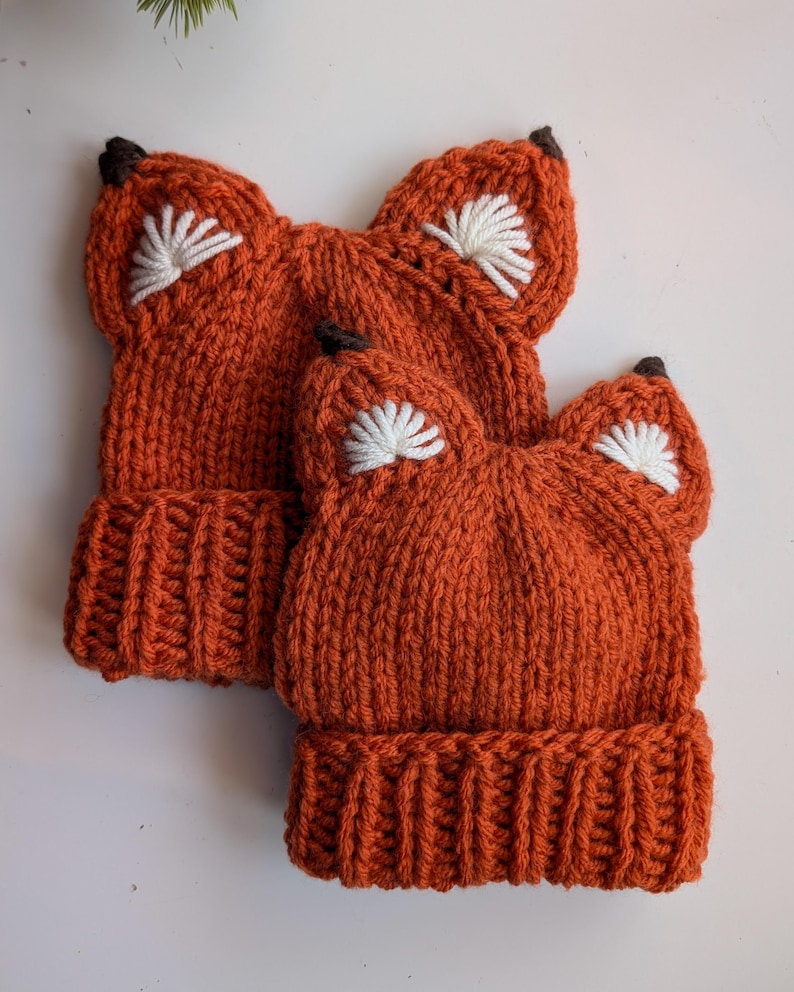 May include: Two orange knitted fox hats with white embroidered ears and brown felt noses.