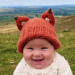 May include: A close-up of a baby wearing a knitted orange fox hat with white accents. The baby is smiling and has rosy cheeks.