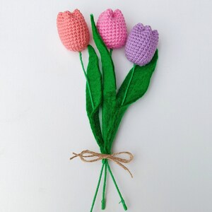 May include: A bouquet of three crocheted tulips in pink, light pink, and purple with green felt leaves tied together with twine.
