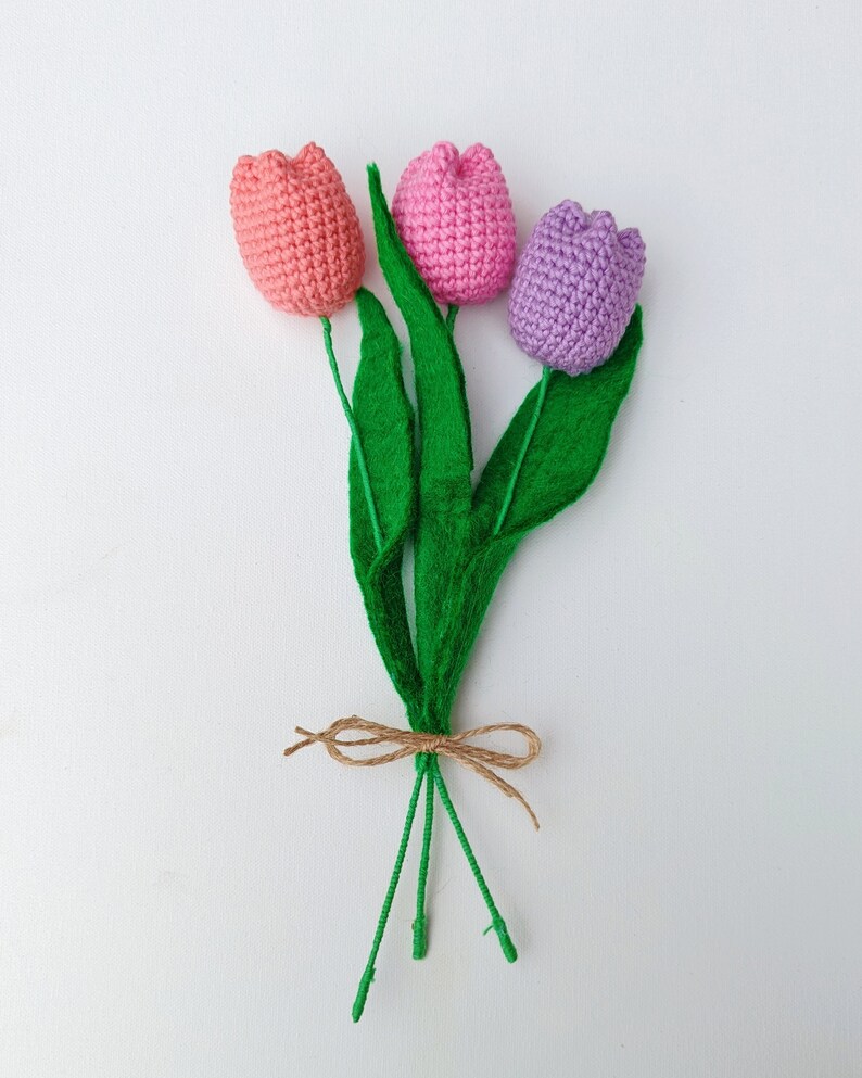 May include: A bouquet of three crocheted tulips in pink, light pink, and purple with green felt leaves tied together with twine.