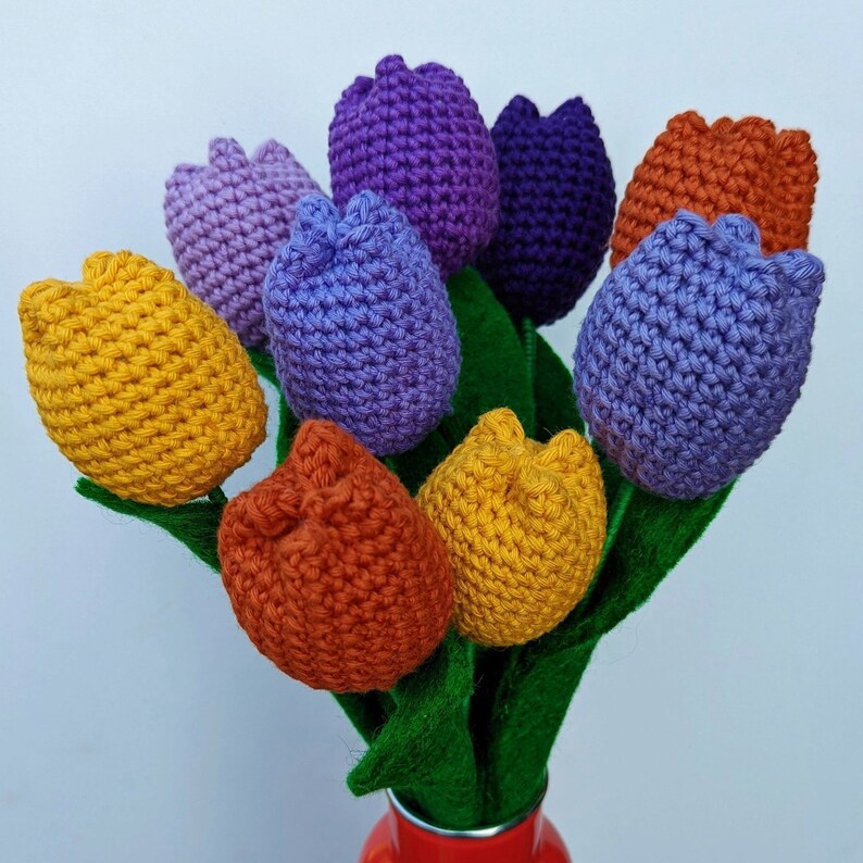 May include: A bouquet of crocheted tulips in various colors, including yellow, orange, purple, and blue. The tulips are arranged in a green felt vase.
