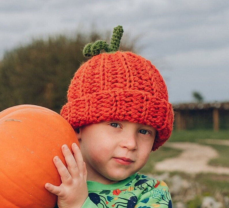 Pode incluir: Uma crian&ccedil;a usando um chap&eacute;u de ab&oacute;bora laranja de croch&ecirc; com um caule verde. A crian&ccedil;a est&aacute; segurando uma ab&oacute;bora laranja grande.