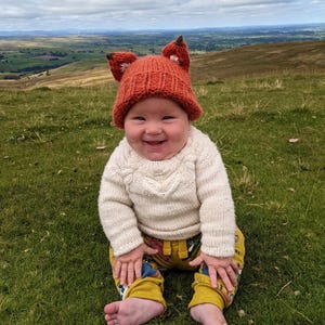 May include: A baby wearing a knitted orange fox hat with white ears and a white sweater sits on a grassy hill. The baby is smiling and looking at the camera.
