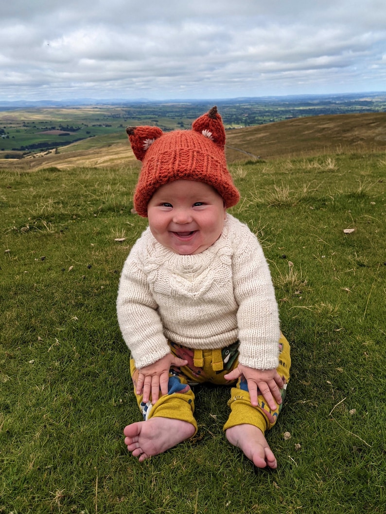 May include: A baby wearing a knitted orange fox hat with white ears and a white sweater sits on a grassy hill. The baby is smiling and looking at the camera.