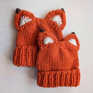 May include: Two orange knitted fox hats with white embroidered ears. The hats are made with a chunky yarn and have a simple, classic design.