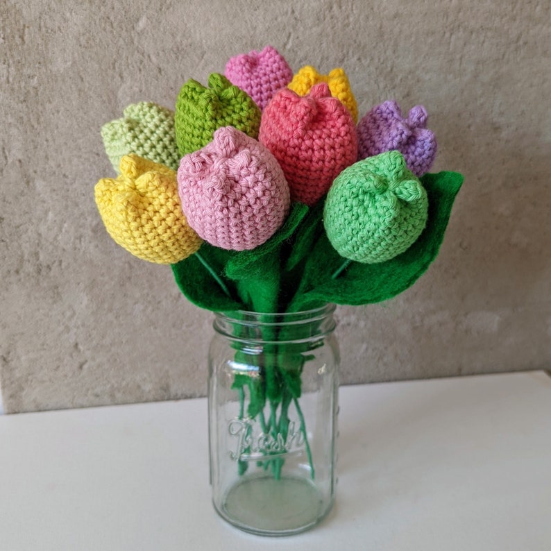 May include: A bouquet of crocheted tulips in a glass jar. The tulips are in shades of pink, yellow, green, and purple.