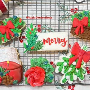 May include: Assortment of decorated Christmas cookies on a wire rack. Designs include wreaths with red bows, a basket of greenery, a candle, a rose, and a cookie with the word "Merry". The text "Whisk Confections" is also visible.