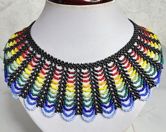 May include: A colorful beaded necklace with a rainbow pattern. The necklace is made of black and multi-colored beads and is displayed on a white mannequin.