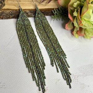 May include: A pair of long, dangling earrings crafted from small, multi-tonal green and metallic seed beads. The earrings feature a triangular top section that transitions into a long, flowing fringe of individual beaded strands hanging from silver-toned hooks.