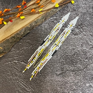 May include: A pair of long, beaded earrings with a white, black, yellow, and brown color scheme. The earrings are hanging from a wooden surface with a floral decoration.
