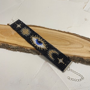 Puede incluir: Pulsera negra de cuentas con un diseño de ojo dorado, blanco y azul, luna y estrellas. La pulsera tiene un cierre de cadena plateada.