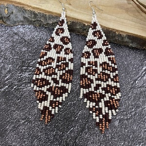 May include: A pair of long, beaded earrings with a brown and white leopard print pattern. The earrings are made of small, round beads and have a teardrop shape.