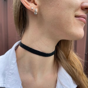 May include: A black choker necklace with a beaded design. The necklace is worn by a person with a white shirt and silver earrings.