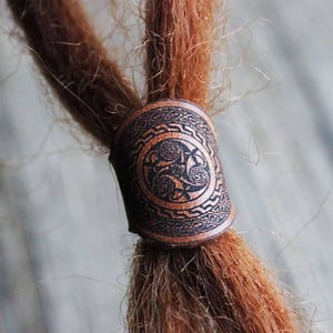 Pode incluir: Um cordão de dreadlocks de couro castanho com um design de nó celta.