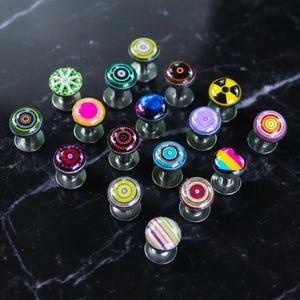 May include: A collection of decorative stud earrings with various colorful, patterned designs. The earrings feature a round, domed top with a metal post and backing. Designs include geometric patterns, swirls, and abstract art.