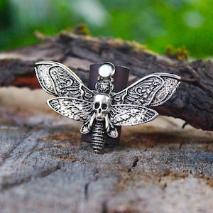Puede incluir: Un anillo plateado con forma de polilla con un diseño de calavera en el cuerpo y patrones detallados en las alas. El anillo está sujeto a una banda de cuero marrón. El anillo está sobre un fondo de madera natural.