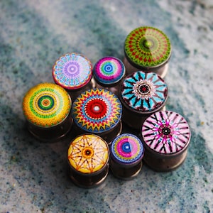 May include: A collection of colorful ear plugs with intricate mandala designs. The plugs feature a variety of colors including red, blue, green, yellow, and purple. The plugs are made of metal and glass.